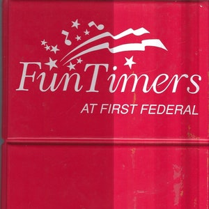 May include: A red plastic sign with the words "Fun Timers at First Federal" in white text. The sign has a white graphic of a flag with stars and musical notes.