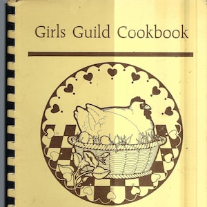 Op de afbeelding: Een geel kookboek met een bruine rand en de titel "Girls Guild Cookbook". De omslag toont een zwart-witte illustratie van een kip in een mand met een geruite achtergrond.