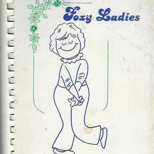 Może przedstawiać: Okładka książki kucharskiej w stylu vintage zatytułowana "Foxy Ladies" z niebieską ilustracją uśmiechniętej kobiety. Okładka zawiera również tekst "An Anthology of Fox Valley Women Cooks by Ellen Kort."