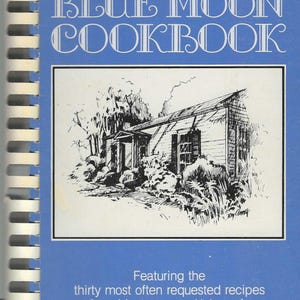 May include: A blue cookbook titled "The Once in a Blue Moon Cookbook." The cover features a black and white illustration of a house. The text below reads "Featuring the thirty most often requested recipes from Montgomery, Alabama's renowned Blue Moon Inn."
