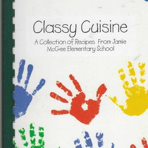 Pode incluir: Um livro de receitas intitulado "Classy Cuisine" com uma capa branca com impressões de mãos coloridas em vermelho, azul, verde e amarelo. O texto diz "A Collection of Recipes From Jamie McGee Elementary School". O livro tem uma lombada verde.