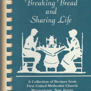 May include: A cookbook cover with the title "Breaking Bread and Sharing Life" in white script on a teal background. The cover features a white silhouette of people seated around a table. The text "A Collection of Recipes from First United Methodist Church Moorestown, New Jersey" is also included.