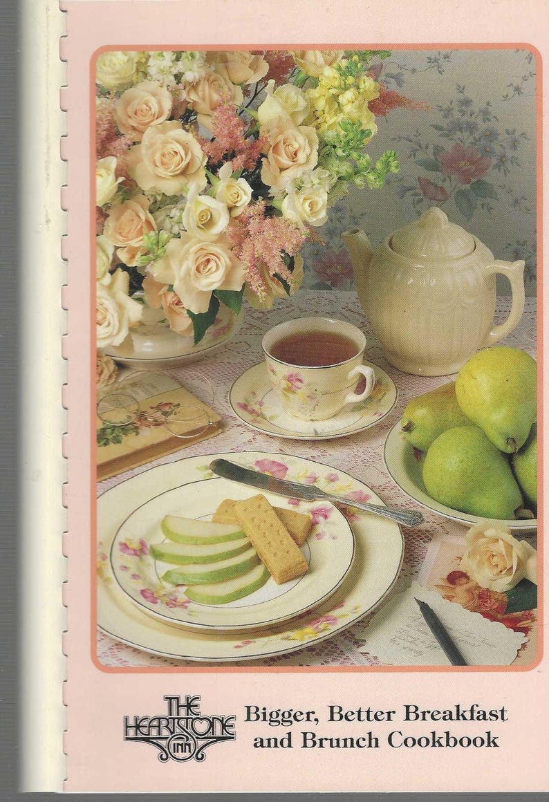 May include: A cookbook cover featuring a breakfast scene. A floral arrangement of roses and other flowers sits near a teapot, teacup, and plates of food, including sliced pears and biscuits. The text reads "Bigger, Better Breakfast and Brunch Cookbook."