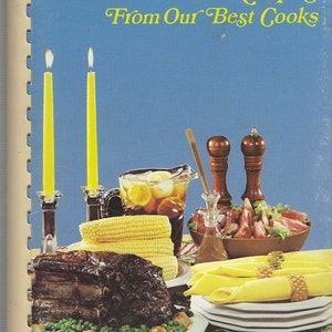 May include: A cookbook titled "Favorite Recipes From Our Best Cooks" with a blue cover and a photo of a table setting with a roast, corn on the cob, a pitcher of iced tea, a salad, and candles.