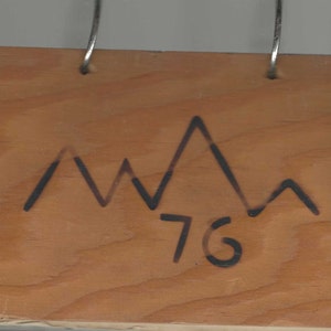 Puede incluir: Un letrero de madera con un dibujo de una cordillera de montañas negras y el número 76 escrito debajo.