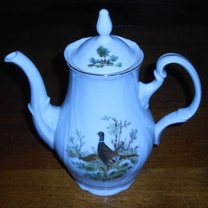 Puede incluir: Tetera de porcelana blanca con ribete dorado y diseño de faisán. La tapa tiene un pequeño diseño de árbol.