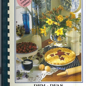 May include: A cookbook titled "DRM - DFAS Treasured Recipes" with a blue and white floral design on the cover. The cover also features a cherry pie, a bowl of cherries, a glass of milk, and a vase of yellow flowers.