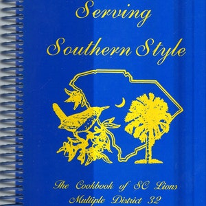 May include: A blue cookbook with a yellow outline of the state of South Carolina, a bird, and a palm tree. The title is "Serving Southern Style" and the text below reads "The Cookbook of SC Lions Multiple District 32".