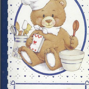 Op de afbeelding: Een witte en blauwe receptenboekomslag met een cartoonbeer die een koksmuts draagt en een kookboek vasthoudt. De tekst op de omslag luidt "Cherished Family Recipes".