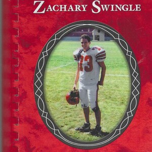 May include: A red cookbook titled "Recipes of Remembrance" with the name "Zachary Swingle" in white text. An oval frame contains a photo of a football player in a white uniform with the number 13. The book is for the Zachary T. Swingle Memorial Scholarship Fund.