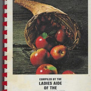 Op de afbeelding: Een vintage kookboekcover met de tekst "Man Shall Not Live By Bread Alone". De afbeelding toont een overvloedshoorn vol rode appels. De cover is gebonden met een rood-witte spiraalbinding.