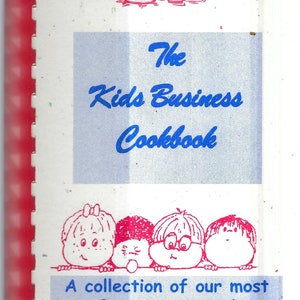 May include: A blue and white cookbook cover with the title "The Kids Business Cookbook" in blue text. The cover features a collection of cartoon children with red hair and pink faces peeking over a white line. The text "A collection of our most favourite recipes!" is in blue text.