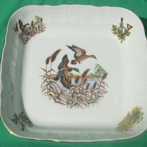 Puede incluir: Plato cuadrado blanco para servir con borde dorado y escena detallada de patos. El centro presenta patos volando sobre juncos y agua. Diseños decorativos en las esquinas con patos, una trompeta y follaje.