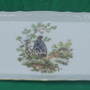 May include: White oval serving platter with a gold rim. The platter features a hand-painted design of birds and foliage in shades of green, brown, and blue.