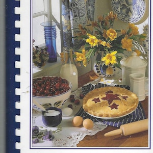 May include: A cookbook titled "Generations of Recipes" with a white background and a blue border. The cover features a still life of a pie, cherries, a milk bottle, eggs, and a rolling pin. The image is set in a kitchen with a window and floral curtains.