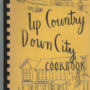 May include: A yellow cookbook with the title "Up Country Down City Cookbook" in black cursive lettering. The cover features a black and white illustration of a small town with buildings and a tree.