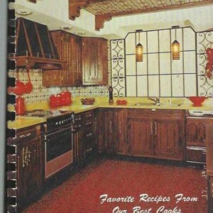 Op de afbeelding: Een vintage kookboek met een rode en witte omslag met de tekst "Favorite Recipes From Our Best Cooks" en "COOK BOOK". De omslag toont een foto van een keuken met houten kasten, een rode tegelvloer en een raam met een smeedijzeren ontwerp.