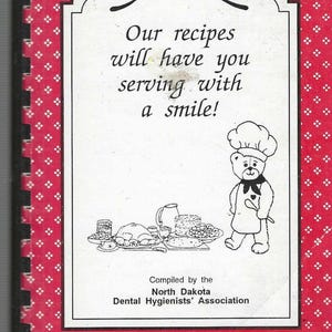 Puede incluir: Una portada de libro de cocina con fondo rojo y marco blanco con el texto "¡Nuestras recetas te harán servir con una sonrisa!". Un oso de dibujos animados chef está junto a ilustraciones de comida. Compilado por la North Dakota Dental Hygienists' Association.