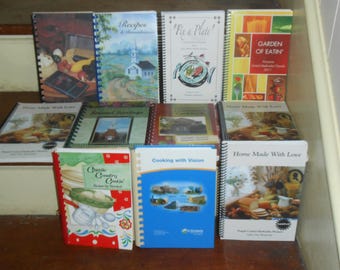 Könnte beinhalten: Eine Sammlung von Kochbüchern mit Spiralbindung und verschiedenen Einbänden. Titel sind u.a. "Recipes", "Fix a Plate", "Garden of Eatin'", "Home Made With Love" und "Cooking with Vision". Die Bücher sind auf einer Holzoberfläche angeordnet.