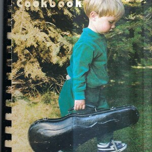 May include: A young boy in a teal sweater holds a black violin case. The book cover behind him reads "The Music Lovers' Cookbook".