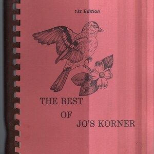 May include: A pink book with a black and white illustration of a bird and a flower. The book title is "The Best of Jo's Korner" and the book is labeled "1st Edition".