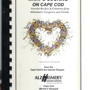 Könnte beinhalten: Ein weißer Buchumschlag mit dem Titel "What's Cooking on Cape Cod" und einem herzförmigen Kranz aus getrockneten Blumen. Das Buch ist eine Sammlung von Lieblingsrezepten und Kommentaren von Alzheimer-Betreuern und Freunden, vom Cape Cod & the Islands Chapter der Alzheimer's Association.