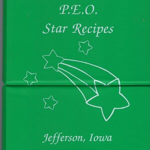 Op de afbeelding: Een groen boekje met de titel "P.E.O. Star Recipes" en de tekst "Jefferson, Iowa" in wit gedrukt. De omslag heeft een witte omtrek van een vallende ster met meerdere strepen.