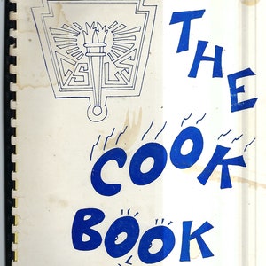 Może przedstawiać: Biała książka kucharska z niebieskim tytułem "THE COOK BOOK". Okładka zawiera czarno-białą ilustrację pochodni i herbu.