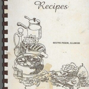 May include: A vintage cookbook titled "Cherished Recipes" with a spiral binding. The cover features a sepia-toned illustration of cooking pots, vegetables, and text that reads "South Pekin, Illinois" and "Lady Funders (ESDA)."