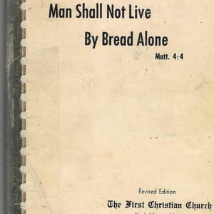 Könnte beinhalten: Ein vergilbtes Buchcover mit dem Titel "Man Shall Not Live By Bread Alone" und der Bibelstelle Matthäus 4,4. Der Einband hat einen cremefarbenen Hintergrund und schwarzen Text. The First Christian Church ist ebenfalls auf dem Cover abgedruckt.