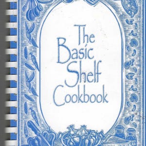 Op de afbeelding: Een spiraalgebonden kookboek getiteld "The Basic Shelf Cookbook" met een blauw-witte omslag. De omslag bevat illustraties van keukengerei, groenten en voedsel, met de titel in elegant schrift.