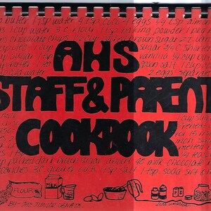 May include: A red and black cookbook cover with the title "AHS Staff & Parent Cookbook". The cover features handwritten recipes for various desserts, including chocolate chip cookies and a chocolate kiss recipe.