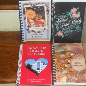 May include: Four spiral-bound cookbooks with various covers. One is red with a heart-shaped cutout, another is black with floral accents, and a third features an American flag design. The fourth cookbook has a brown cover.