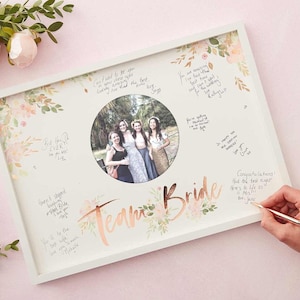 Könnte beinhalten: Ein weißer Rahmen mit floralem Design und einem Foto einer Gruppe von Freundinnen. Der Rahmen trägt die Aufschrift "Team Bride" in goldenen Lettern. Um das Foto herum befinden sich handschriftliche Nachrichten und die Worte "Congratulations! Had the best night! Here's to life as Mrs. Jane" sind in der rechten unteren Ecke geschrieben.