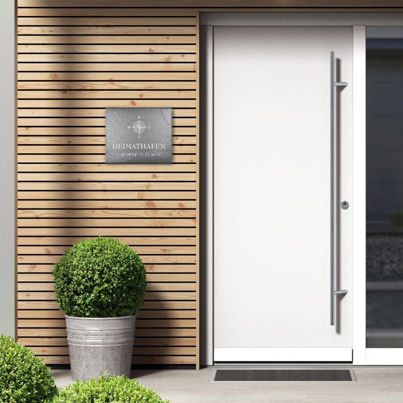 May include: A gray slate sign with a compass and the text "HEIMATHAFEN N 48&deg;18'12" O 11&deg;34'31"". The sign is mounted on a wooden wall with a white door to the right.