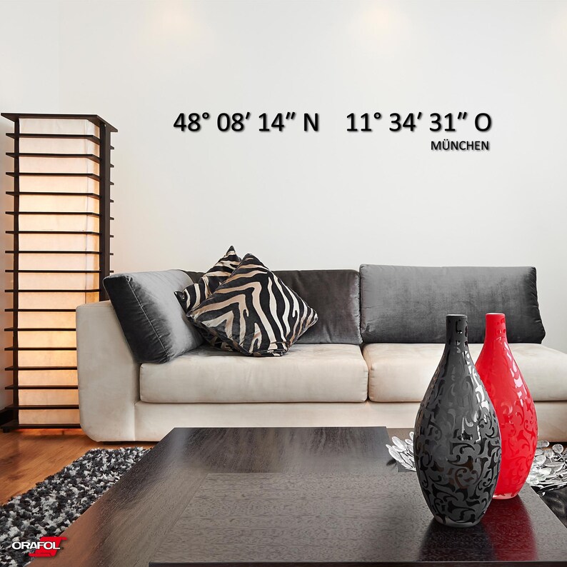 May include: A white couch with gray and zebra print throw pillows sits in a living room with a wooden floor. Two decorative vases, one black and one red, are on a black coffee table in front of the couch. The wall behind the couch has the coordinates 48&deg; 08&prime; 14" N 11&deg; 34' 31" O and the word M&Uuml;NCHEN written below.