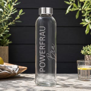 Könnte beinhalten: Eine klare Glaswasserflasche mit einem silberfarbenen Metalldeckel. Die Flasche ist mit vertikal gedrucktem weißem Text versehen, der POWERFRAU und den Namen Bea in einer Schreibschrift zeigt. Das Design ist mittig auf der Flasche platziert.