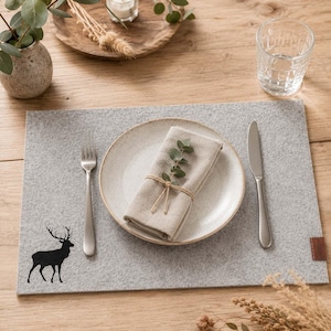 Può includere: Un sottopiatto in feltro grigio con una silhouette di cervo nero, un piatto beige, un tovagliolo di lino piegato legato con spago e posate. L'apparecchiatura include un bicchiere d'acqua e un vassoio di legno con una candela e verde.