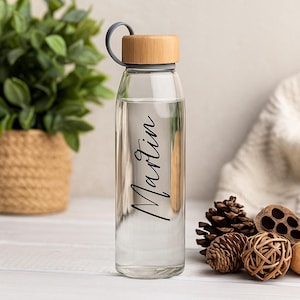 Könnte beinhalten: Eine transparente Glaswasserflasche mit Holzdeckel und dem Namen "Martin" in schwarzer Schrift. Die Flasche hat einen grauen Griff und ist mit Wasser gefüllt. Die Flasche ist etwa 23 cm hoch.