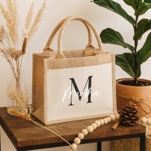 Könnte beinhalten: Beige Jute-Tragetasche mit cremefarbener Frontplatte. Auf der Platte ist ein großer schwarzer Buchstabe "M" und der Name "Martha" in Schreibschrift zu sehen. Die Tasche hat passende beige Griffe und Verzierungen.