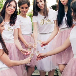 Könnte beinhalten: Fünf Freundinnen tragen weiße T-Shirts mit goldenen Schriftzügen. Die Shirts sagen "Team Bride" und "Bride Charlotte". Sie halten Sektgläser in der Hand und tragen rosa Tüllröcke.