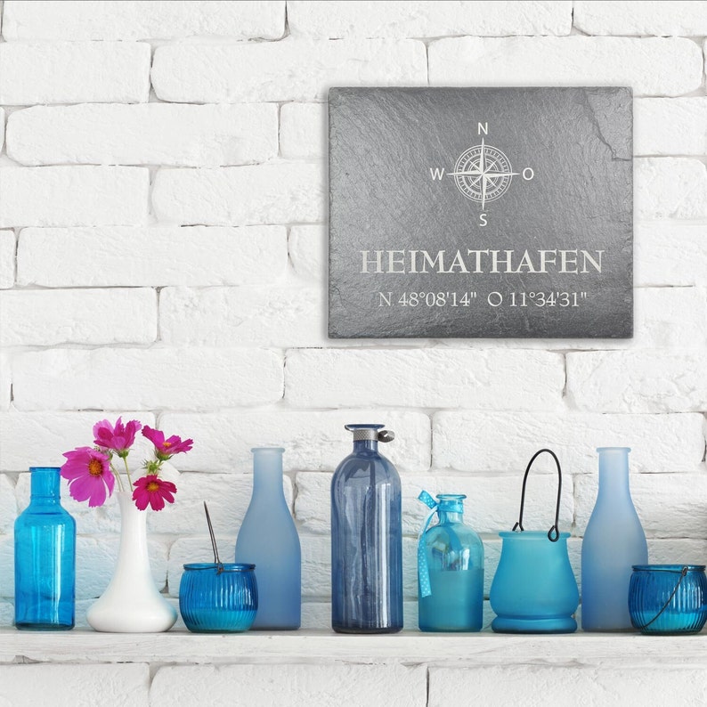 May include: A gray slate sign with a compass rose and the text "HEIMATHAFEN N 48'08'14" O 11'34'31". The sign is hanging on a white brick wall. There are blue glass bottles and a vase with pink flowers on a white shelf below the sign.