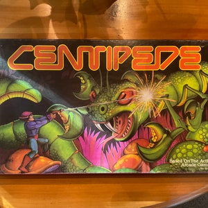May include: A board game box with a green centipede illustration on the front. The box is red, black, and white with the title "CENTIPEDE" in red letters. The text "Based On The Action-Packed Arcade Game By ATARI." is at the bottom of the box.