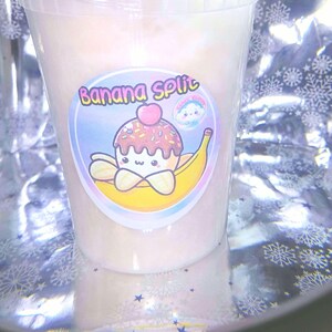 Puede incluir: Un vaso de plástico transparente lleno de una sustancia rosa claro, posiblemente un postre. El vaso tiene una tapa y una pegatina que dice "Banana Split" con un helado de dibujos animados sobre un plátano. El fondo es brillante.