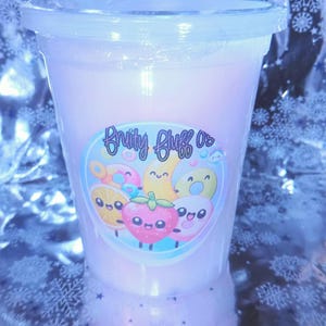 May include: A clear plastic cup filled with pink cotton candy, sealed with a clear lid. A colorful sticker on the cup features cartoon fruit characters and the text "Fruity Bluffs Os". The background has a snowflake pattern.