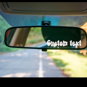 May include: A black rearview mirror with the text "Custom text" in white, set against a blurred background of a road and trees. The mirror is mounted on a car's windshield.