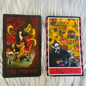 May include: Two tarot cards featuring gothic illustrations. One card depicts a woman in a black dress on a red chair, labeled "The Empress." The other card shows a figure in a Scream mask with the text "What's your favorite scary movie?" and "Hello Sidney," labeled "The Fool."