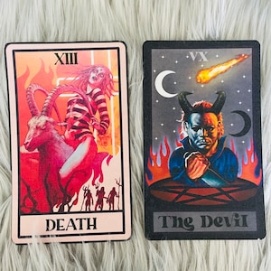 May include: Two tarot cards with dark backgrounds and illustrations. One card features a figure riding a goat with the word "DEATH". The other card depicts a devilish figure with the words "The Devil". Both cards have a black border.