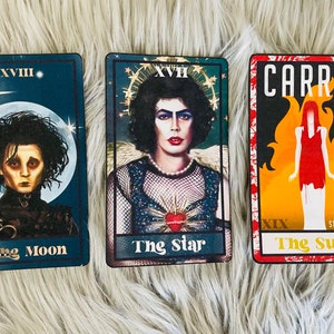 May include: Three tarot cards featuring iconic characters. "The Moon" card shows Edward Scissorhands. "The Star" card features a character in a mesh top and pearls. "The Sun" card has the title "Carrie" and a figure in a red dress.