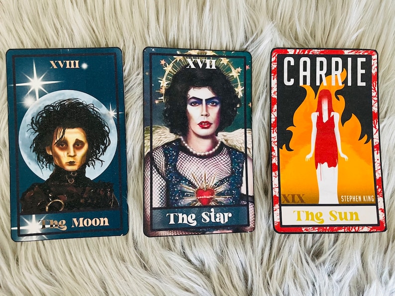 May include: Three tarot cards featuring iconic characters. "The Moon" card shows Edward Scissorhands. "The Star" card features a character in a mesh top and pearls. "The Sun" card has the title "Carrie" and a figure in a red dress.